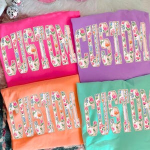 Puede incluir: Cuatro camisetas coloridas: rosa, morado, naranja y verde azulado. Cada camiseta presenta la palabra "CUSTOM" en un dise&ntilde;o floral. Las letras est&aacute;n delineadas en blanco con un borde perlado. El dise&ntilde;o floral incluye flores rosas, naranjas y azules.