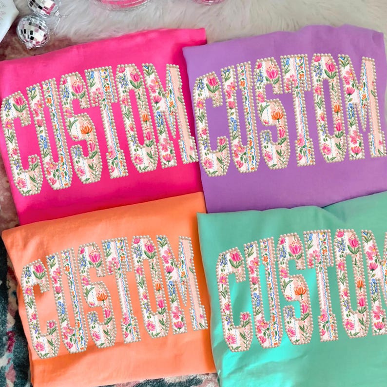 Puede incluir: Cuatro camisetas coloridas: rosa, morado, naranja y verde azulado. Cada camiseta presenta la palabra "CUSTOM" en un dise&ntilde;o floral. Las letras est&aacute;n delineadas en blanco con un borde perlado. El dise&ntilde;o floral incluye flores rosas, naranjas y azules.