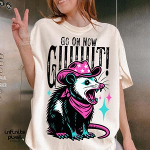 May include: Cream-colored t-shirt featuring a cartoon opossum wearing a pink cowboy hat with white stars and a matching bandana. The opossum is screaming, with the words "GO ON NOW" and "GIT IT!" in black text.