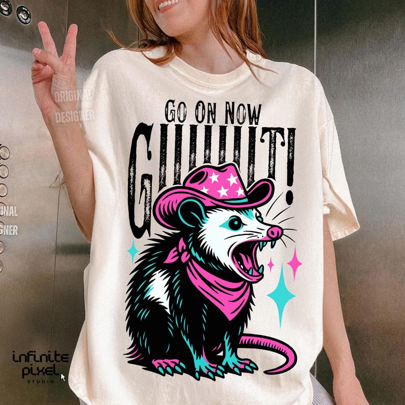 May include: Cream-colored t-shirt featuring a cartoon opossum wearing a pink cowboy hat with white stars and a matching bandana. The opossum is screaming, with the words "GO ON NOW" and "GIT IT!" in black text.