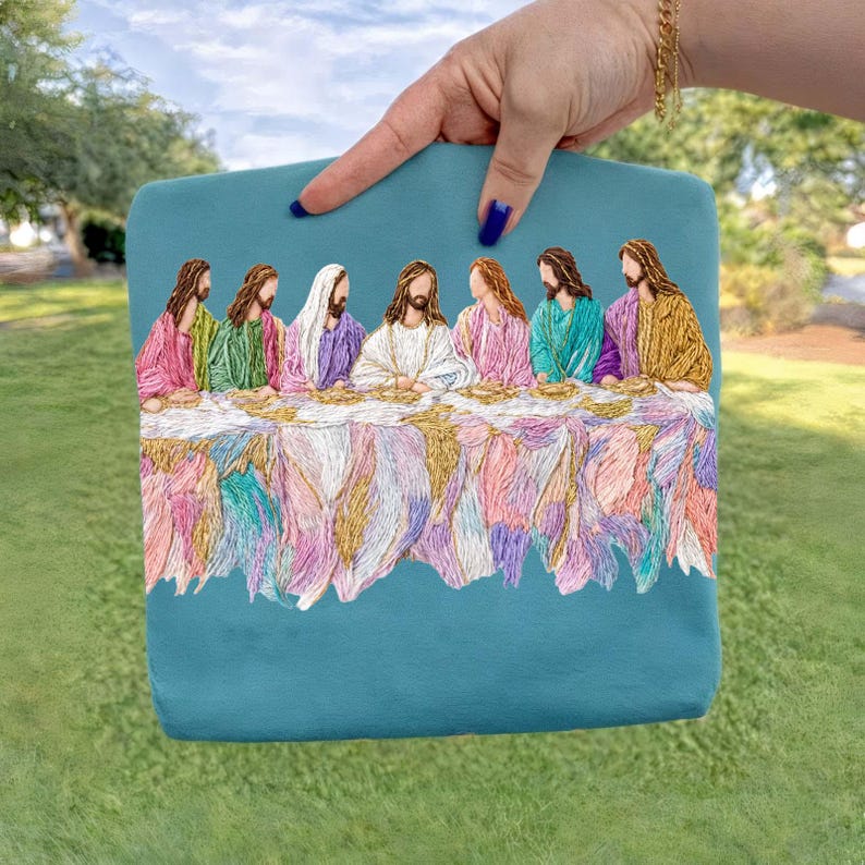 May include: Teal-colored embroidered clutch featuring a colorful depiction of the Last Supper. The embroidery showcases Jesus and his disciples with detailed thread work in various colors. The clutch is held by a hand with blue nail polish.