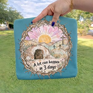 May include: Teal tote bag featuring a watercolor design of a sunrise over a landscape with a tomb and crosses, framed by a crown of thorns. The text "A lot can happen in 3 days" is printed below the image.
