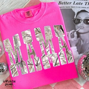 May include: A bright pink crew neck sweatshirt with the word "MAMA" in large, cutout letters filled with a camouflage pattern. The shirt has a tag that says "Comfort Colors". A disco ball and a black and white image are in the background.