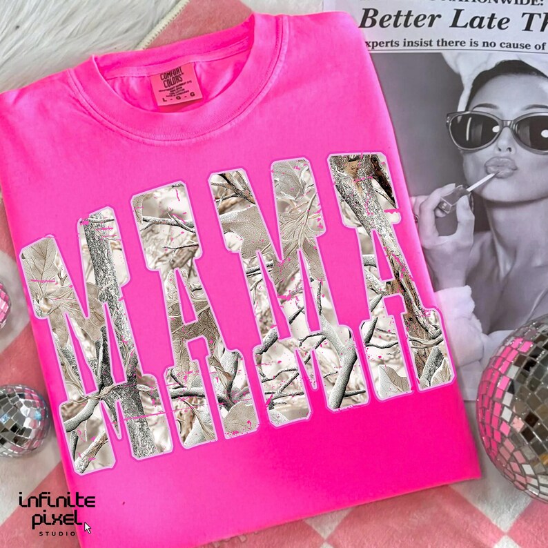 May include: A bright pink crew neck sweatshirt with the word "MAMA" in large, cutout letters filled with a camouflage pattern. The shirt has a tag that says "Comfort Colors". A disco ball and a black and white image are in the background.