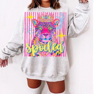 May include: A light gray sweatshirt featuring a colorful leopard illustration wearing a crown and necklace. The leopard is set against a pink striped background with the word "Spoiled" in yellow script.