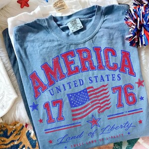 May include: A light blue t-shirt with the word "AMERICA" in red block letters, along with "UNITED STATES", "1776", and a flag graphic. The shirt is XL size.