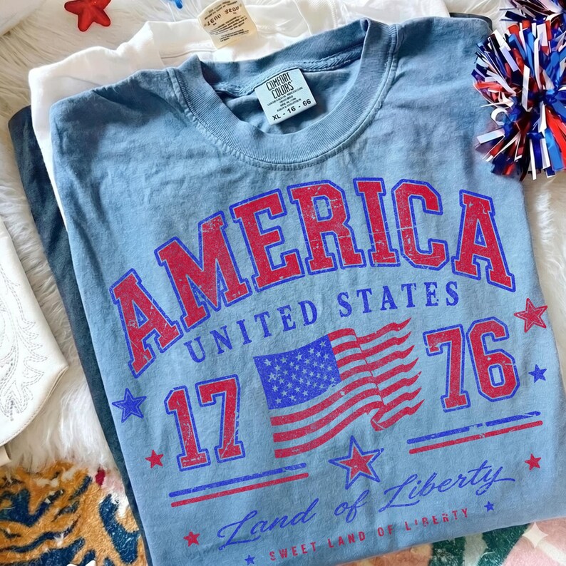 May include: A light blue t-shirt with the word "AMERICA" in red block letters, along with "UNITED STATES", "1776", and a flag graphic. The shirt is XL size.