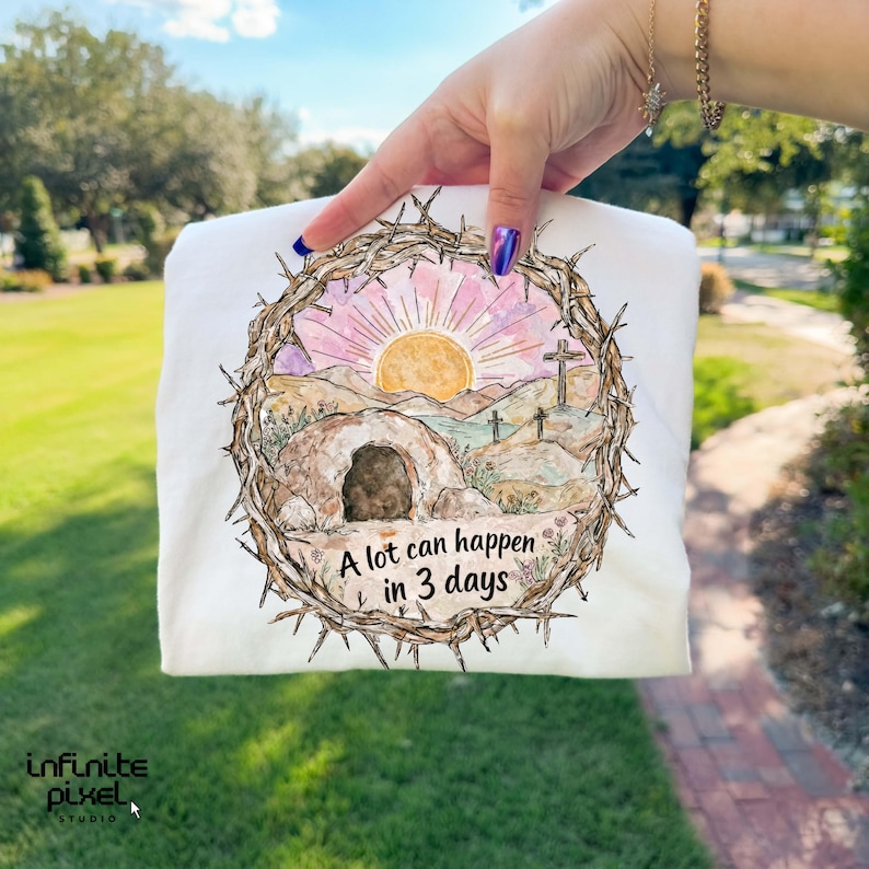 May include: White sweatshirt featuring a watercolor design of a sunrise over a landscape with a tomb and crosses, framed by a crown of thorns. The text "A lot can happen in 3 days" is below the image.