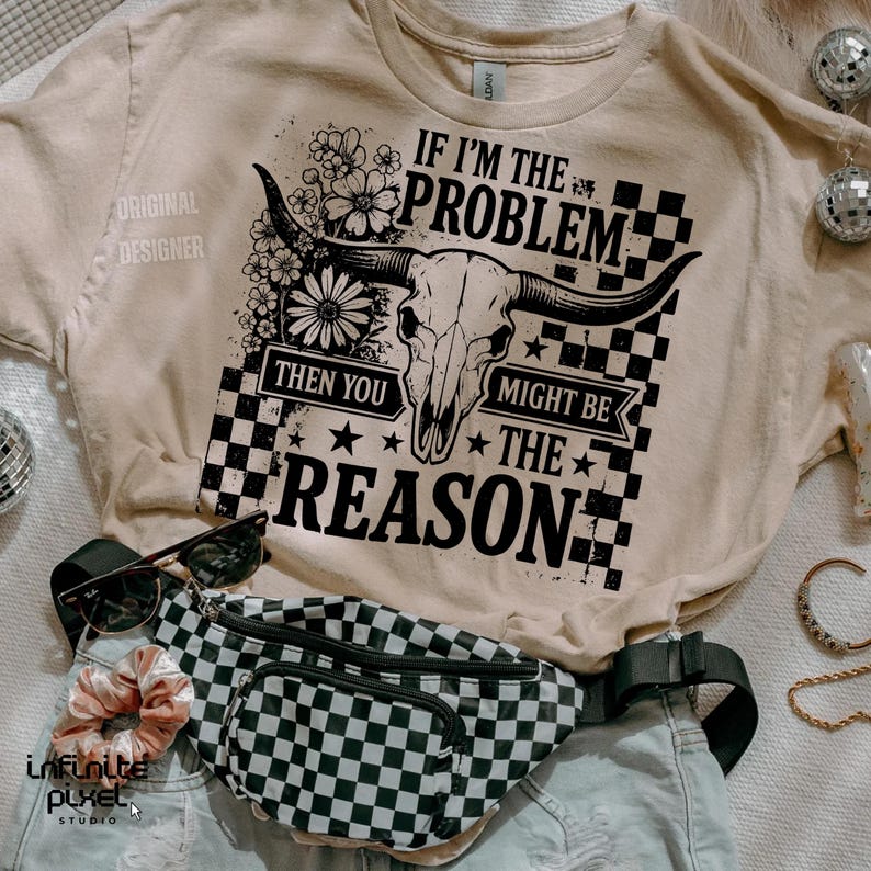 May include: Beige t-shirt with a graphic featuring a bull skull, flowers, and the text "IF I'M THE PROBLEM THEN YOU MIGHT BE THE REASON." A black and white checkered fanny pack, sunglasses, and a scrunchie are also visible.