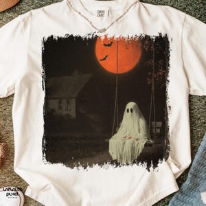 May include: White t-shirt featuring a spooky Halloween design. The graphic depicts a ghost on a swing set under a large orange moon, with bats and a house in the background. The shirt has a Comfort Colors tag.