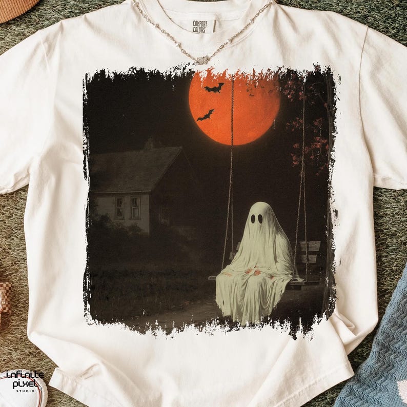 May include: White t-shirt featuring a spooky Halloween design. The graphic depicts a ghost on a swing set under a large orange moon, with bats and a house in the background. The shirt has a Comfort Colors tag.