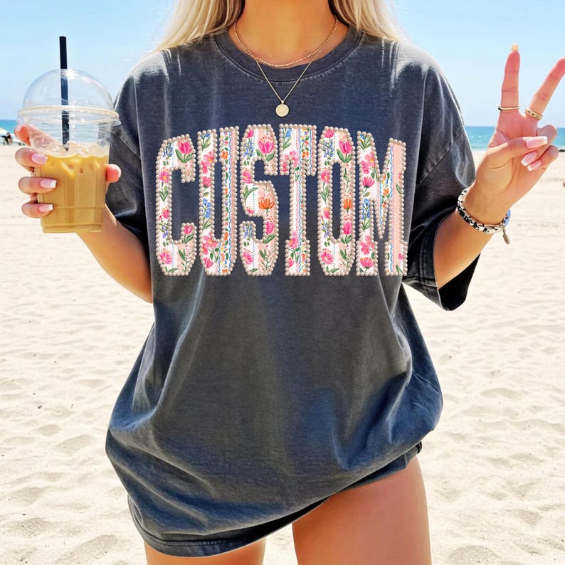 Puede incluir: Camiseta gris oscuro con la palabra "CUSTOM" en un dise&ntilde;o floral. Las letras est&aacute;n llenas de flores rosas, azules y blancas. La camiseta se usa en una playa.