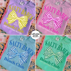 May include: Four t-shirts in pastel colors: lavender, pink, blue, and green. Each shirt features the text "SALTY BABES" above a bow graphic and "Beach Club Est. 2025 Girls Just Wanna Have Sun" below. The bow and text colors match the shirt colors.