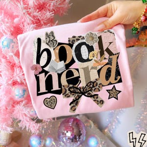 May include: A light pink t-shirt with the words "book nerd" in a collage style design. The design includes a butterfly, open book, bow, and star. The shirt is held up by a person's hand.
