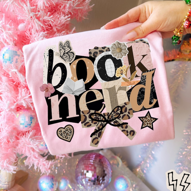 May include: A light pink t-shirt with the words "book nerd" in a collage style design. The design includes a butterfly, open book, bow, and star. The shirt is held up by a person's hand.