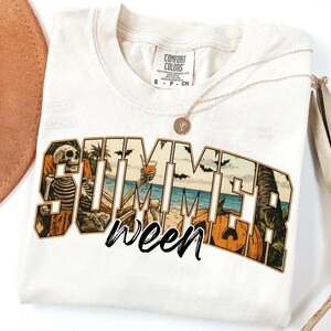 May include: Cream-colored t-shirt with a graphic design featuring the word "SUMMER" with a beach scene and a skeleton. The word "ween" is written below in a script font. A gold necklace is draped across the shirt.
