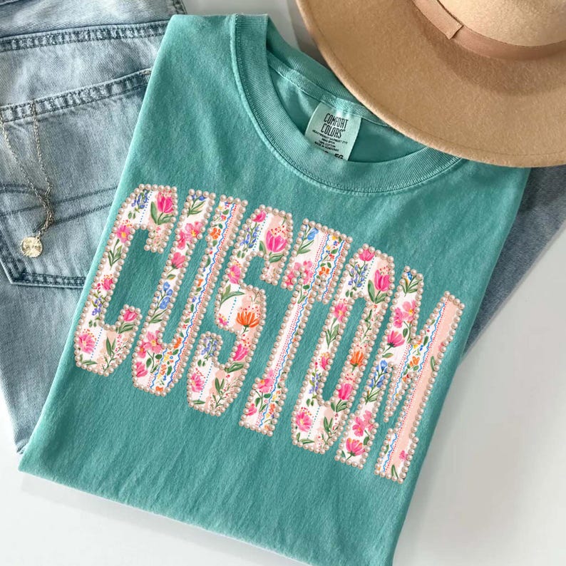 Puede incluir: Una camiseta Comfort Colors turquesa con la palabra "CUSTOM" en un dise&ntilde;o floral. Las letras est&aacute;n rellenas de flores rosas, naranjas y azules. La camiseta est&aacute; doblada sobre unos vaqueros azules y un sombrero beige.