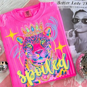 May include: Bright pink t-shirt featuring a colorful leopard illustration wearing a jeweled crown and necklace. The word "SPOILED" is written in yellow script. The shirt has a vintage, washed look.