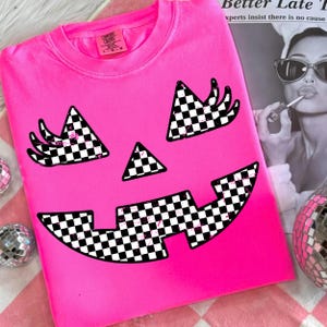 May include: A bright pink Comfort Colors sweatshirt featuring a jack-o'-lantern face design. The eyes, nose, and mouth are black and white checkered patterns. The shirt is folded and displayed on a pink and white striped surface.