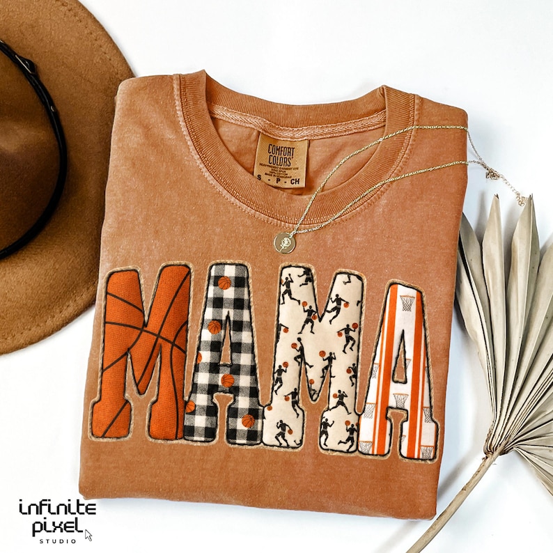 Puede incluir: Una camiseta Comfort Colors de color naranja quemado con la palabra "MAMA" en letras grandes y decorativas. La "M" presenta un dise&ntilde;o de baloncesto, la "A" es un patr&oacute;n de cuadros blancos y negros, la segunda "M" tiene siluetas de jugadores de baloncesto y la "A" final tiene un dise&ntilde;o de aro de baloncesto.