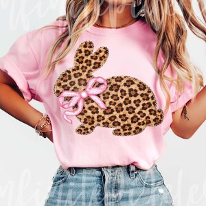 May include: A pink t-shirt with a leopard print bunny design and a pink bow. The bunny has a stitched outline. The shirt is made of a soft material, suitable for casual wear. The design is centered on the front of the shirt.
