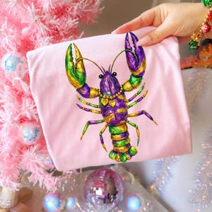 May include: A light pink t-shirt featuring a colorful illustration of a lobster. The lobster is decorated with purple, green, and gold accents, and a gold necklace with heart pendants. The design is vibrant and eye-catching.