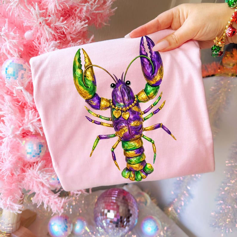 May include: A light pink t-shirt featuring a colorful illustration of a lobster. The lobster is decorated with purple, green, and gold accents, and a gold necklace with heart pendants. The design is vibrant and eye-catching.