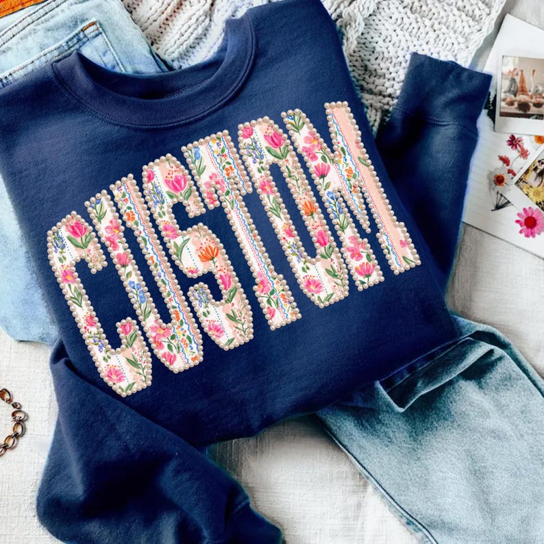 Puede incluir: Sudadera azul marino con la palabra "CUSTOM" en un dise&ntilde;o floral. Las letras est&aacute;n rellenas de flores rosas, naranjas y azules, delineadas con un ribete perlado. La sudadera est&aacute; doblada sobre unos vaqueros azules.