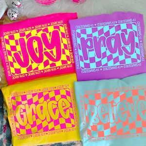 May include: Four colorful t-shirts with the words "JOY", "PRAY", "GRACE", and "BELIEVE" in bold, retro fonts. Each shirt features a checkered design in contrasting colors and Bible verses. The shirts are in pink, purple, yellow, and blue.