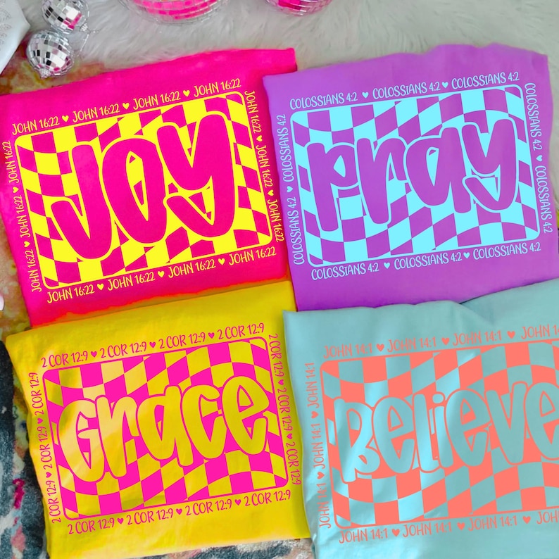May include: Four colorful t-shirts with the words "JOY", "PRAY", "GRACE", and "BELIEVE" in bold, retro fonts. Each shirt features a checkered design in contrasting colors and Bible verses. The shirts are in pink, purple, yellow, and blue.
