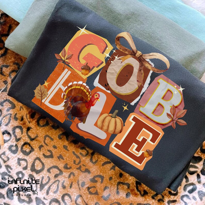 May include: A dark gray t-shirt with the word "GOBBLE" in colorful, block letters. The design includes a turkey, pumpkin, and fall leaves. The shirt is folded on a leopard print surface.