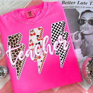 May include: A bright pink sweatshirt with the word "teacher" in white script. The word is overlaid on three lightning bolt designs with leopard print, cherry, and checkered patterns. The shirt has a crew neck and long sleeves.