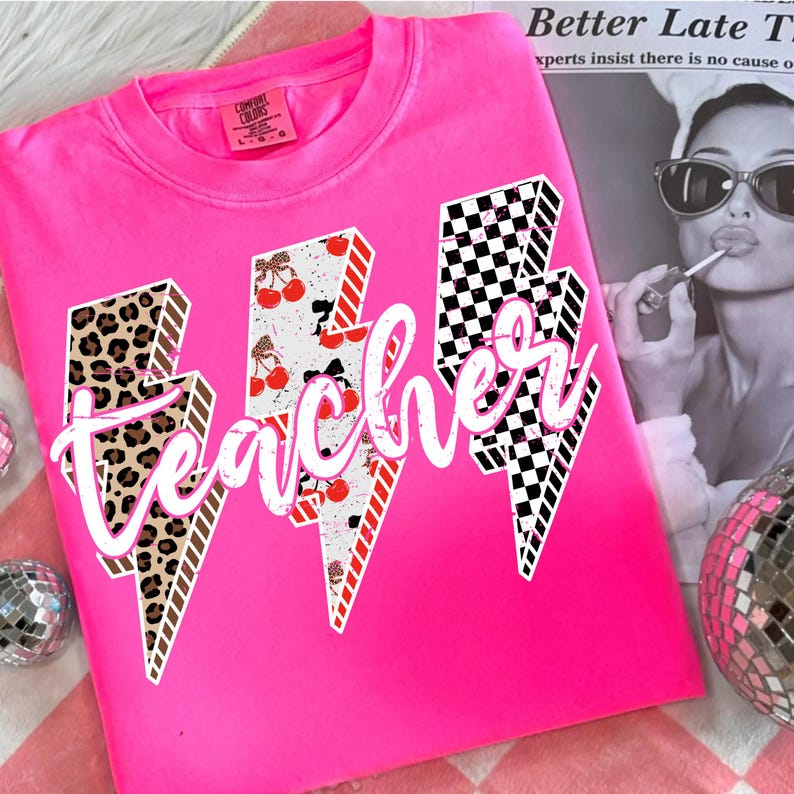 May include: A bright pink sweatshirt with the word "teacher" in white script. The word is overlaid on three lightning bolt designs with leopard print, cherry, and checkered patterns. The shirt has a crew neck and long sleeves.