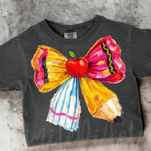 May include: Dark gray t-shirt featuring a colorful bow design. The bow is made of a yellow pencil, a red apple, and a pink, yellow, and black ribbon. The bow has blue and white striped accents. The shirt has the words "Comfort Colors" on the tag.