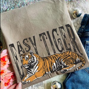 May include: A tan t-shirt with a reclining tiger illustration in orange and black stripes. The text "EASY TIGER" is printed above the tiger in a black, bold font. The shirt is folded and placed on a surface with denim jeans and decorative elements.