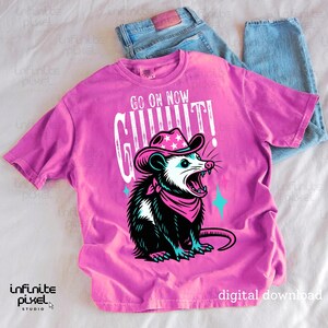 May include: A pink t-shirt with a graphic of a possum wearing a cowboy hat and bandana, with the text "GO ON NOW GIT!" in white. The shirt is paired with blue jeans.