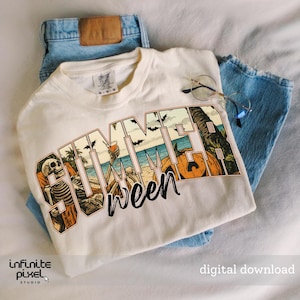 May include: Cream-colored t-shirt with the word "SUMMERween" in a beach-themed graphic. The design includes skeletons, pumpkins, palm trees, and bats. The shirt is laid on top of blue jeans, with a pair of glasses resting on the shirt.