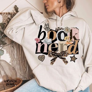 May include: Beige hoodie with the words "book nerd" in a collage of fonts and patterns. The design includes a butterfly, flowers, a heart, and a star. The hoodie is a casual style.