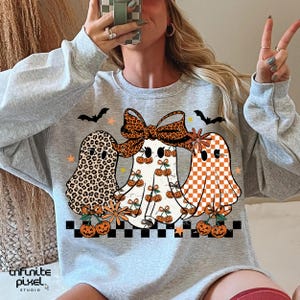 May include: A light gray sweatshirt featuring three cartoon ghosts with Halloween-themed designs. One ghost has a leopard print, another has cherries, and the third has an orange and white checkerboard pattern. Bats, stars, and pumpkins complete the design.