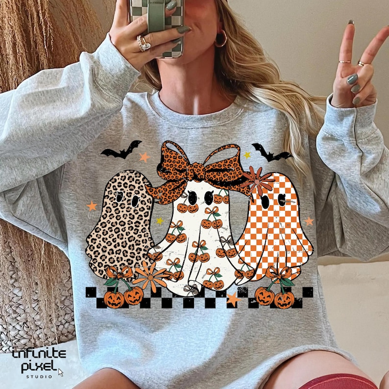 May include: A light gray sweatshirt featuring three cartoon ghosts with Halloween-themed designs. One ghost has a leopard print, another has cherries, and the third has an orange and white checkerboard pattern. Bats, stars, and pumpkins complete the design.