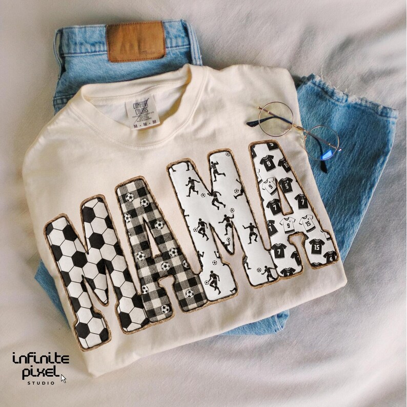 May include: Cream-colored sweatshirt with the word "MAMA" in large, decorative letters. The "M" and "A" are patterned with soccer balls, the second "A" features a black and white checkered pattern, and the last "A" has soccer player silhouettes and jerseys. Blue jeans and glasses are in the background.