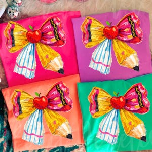 May include: Four colorful t-shirts, each featuring a unique design. The design is a pencil-shaped bow with a red apple in the center. The shirts are in pink, purple, orange, and green.