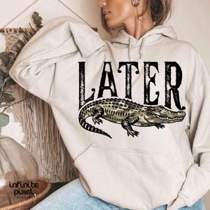 Peut inclure: Un sweat &agrave; capuche beige avec le mot "LATER" en noir et un motif d'alligator. L'alligator est d&eacute;taill&eacute; en vert, gris et marron. Le sweat &agrave; capuche a une poche avant et une capuche avec cordon de serrage.