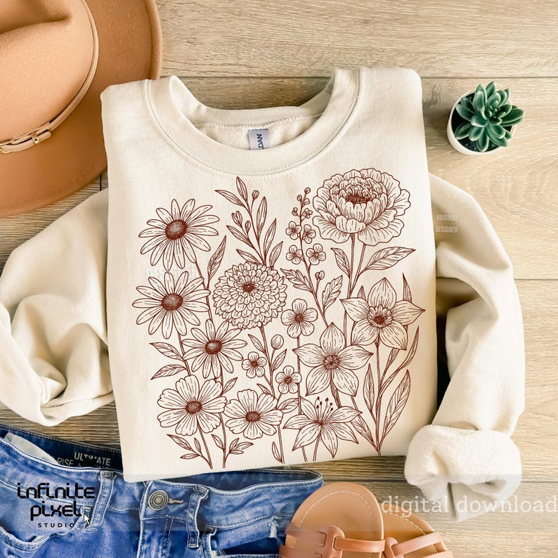 May include: Cream-colored sweatshirt featuring a detailed brown floral design. The design includes various flowers and foliage, creating a botanical illustration. The sweatshirt is laid on top of blue jeans, a hat, and sandals.