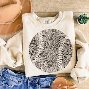 May include: Cream-colored sweatshirt featuring a baseball design with a leopard print pattern. The baseball graphic is centered on the front. The image also includes a tan hat, blue jeans, and sandals, suggesting a casual, sporty theme.