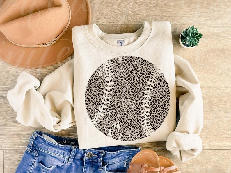 May include: Cream-colored sweatshirt featuring a baseball design with a leopard print pattern. The baseball graphic is centered on the front. The image also includes a tan hat, blue jeans, and sandals, suggesting a casual, sporty theme.