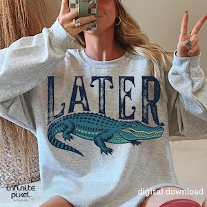 Pode incluir: Camisola cinzenta com a palavra "LATER" impressa em preto e um gr&aacute;fico de um jacar&eacute; azul.