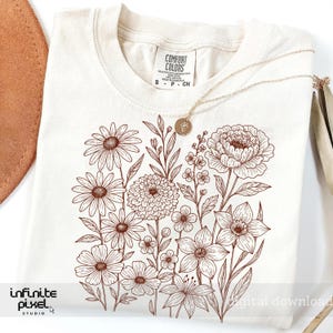 May include: Cream-colored t-shirt with a brown floral design featuring various flowers and foliage. A gold necklace with a pendant rests on the shirt. The shirt has a tag that reads "Comfort Colors".
