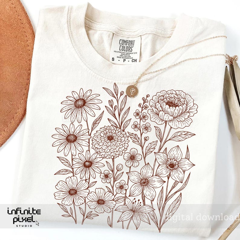 May include: Cream-colored t-shirt with a brown floral design featuring various flowers and foliage. A gold necklace with a pendant rests on the shirt. The shirt has a tag that reads "Comfort Colors".