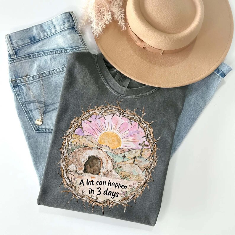 May include: A gray t-shirt with a watercolor graphic of a sunrise over a landscape with crosses and the text "A lot can happen in 3 days." The shirt is paired with light blue jeans and a tan hat.